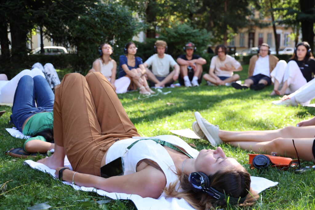 A group of adults sit in a circle in the grass, and a few lie down, all with their eyes closed in a meditation session.