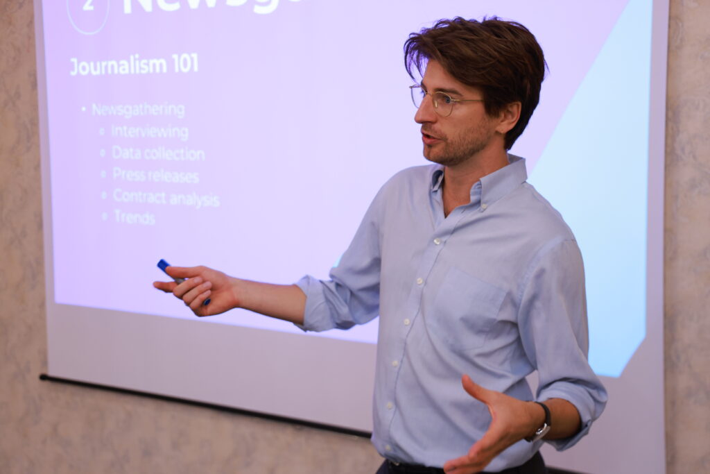 An adult stands in front of a projector screen that says "Journalism 101" with bullet points under it that say "Interviewing, data collection, press releases, contract analysis, trends."