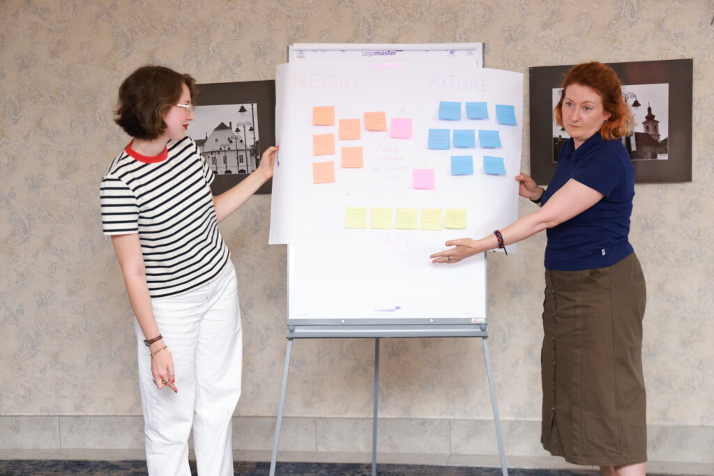 Two adults stand next to a whiteboard with sticky notes arranged around it.