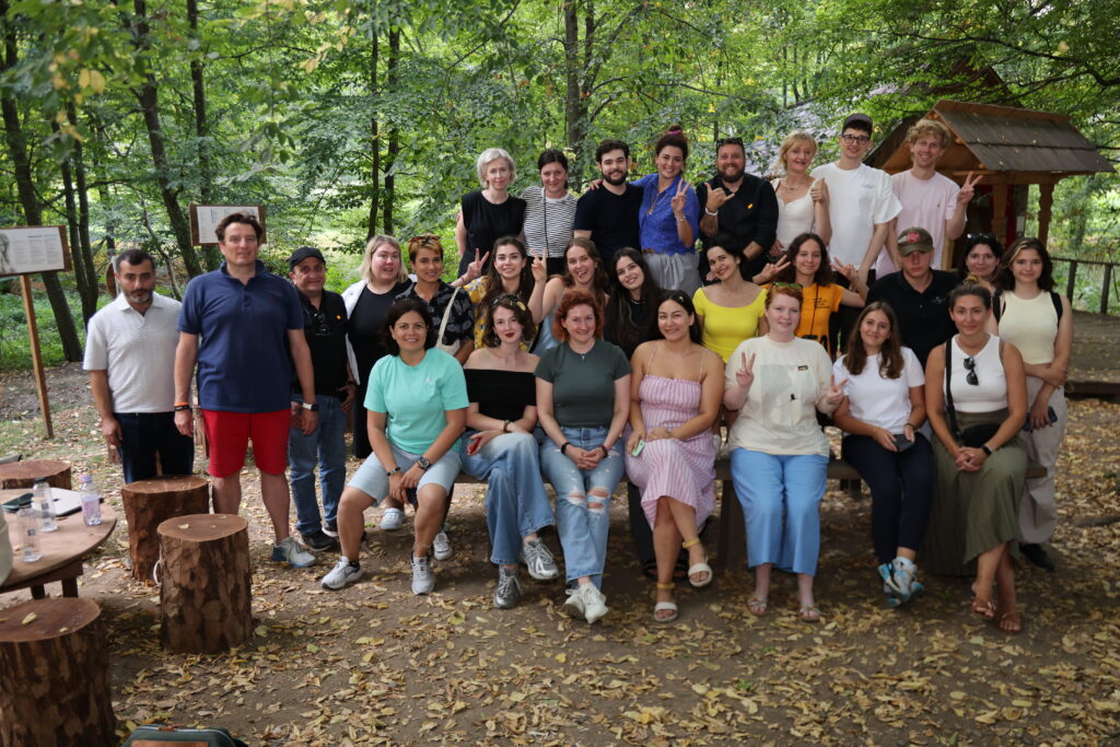 Twenty-eight adults pose for a group photo at a woodsy location.