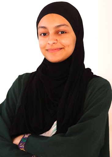 Young woman wearing a black hijab has her arms folded and is smiling toward the camera ahead.