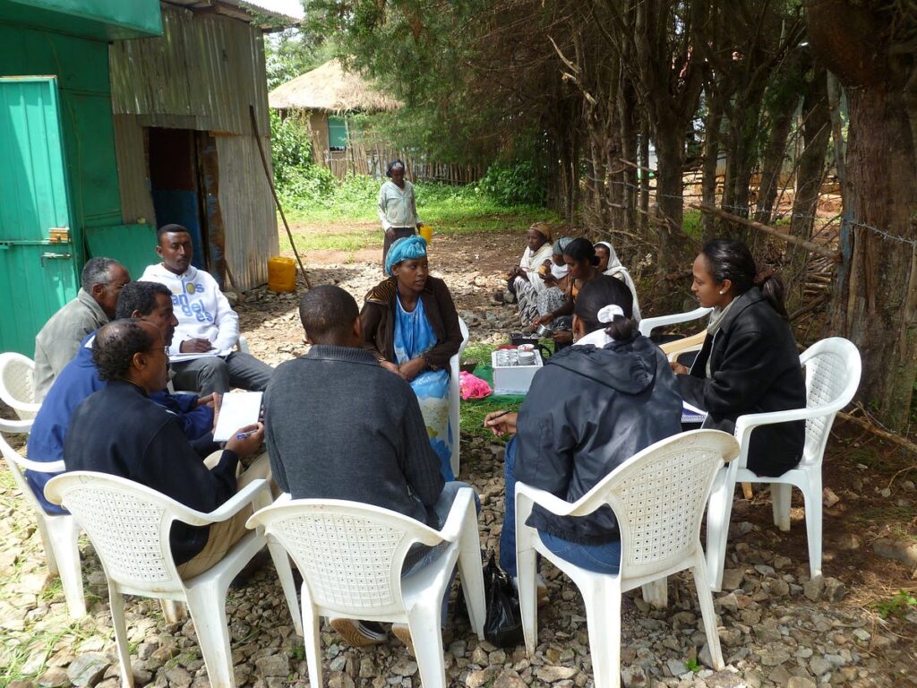 Nine adults sit in an outdoor location in white chairs formed in a circle. Three additional adults are sitting in a row off to the side, while another walks in the distance.