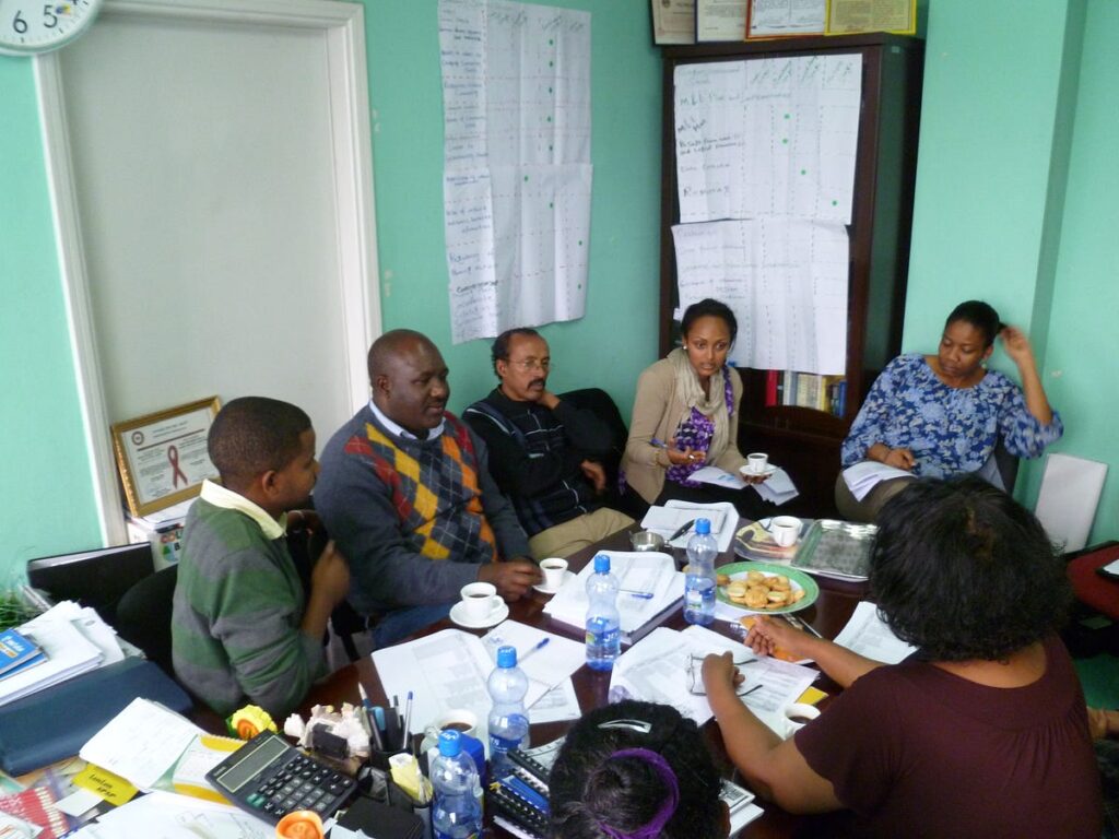 Six adults sit around a table in an indoor office setting. On the table are scattered papers, plates of food, and water bottles.