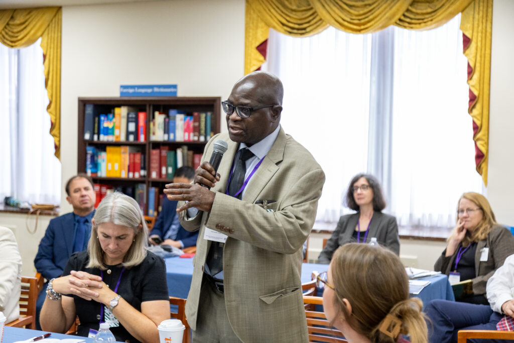 An adult stands and speaks into a microphone in a room of symposium participants, who are sitting down at a table around him.