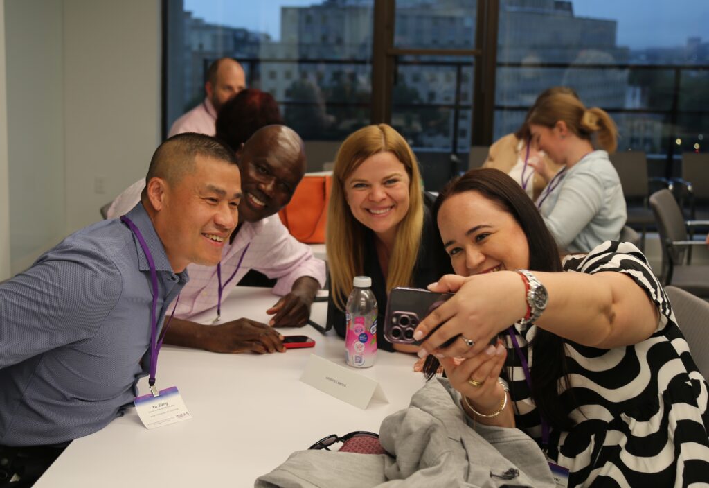 Four adults pose for a group selfie together at a table. Four other adults can be seen in the background in front of a window.